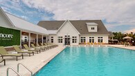 Located at 2626 Goose Island Drive in Leland, North Carolina, this single-story community amenity center at Encore at Leland is designed around a resort-inspired outdoor experience. At its centerpiece is a large, sparkling swimming pool with vivid blue water, framed by a spacious concrete sun deck lined with cushioned lounge chairs and tables — ideal for relaxing in the coastal Carolina climate.

The clubhouse building is architecturally styled in a modern farmhouse aesthetic, featuring crisp white board-and-batten siding, a steeply pitched roofline with a combination of standing seam metal and asphalt shingle roofing, and large multi-pane windows that allow abundant natural light into the interior. A covered outdoor pavilion extends from the main structure, providing a shaded gathering area equipped with additional seating and a large branded green wall displaying the Encore community name.

The pool deck is accessible via ADA-compliant entry equipment and includes water features that add a dynamic element to the pool experience. Surrounding the facility, views of the broader Encore residential community — with its palm trees and multi-story homes — reinforce the neighborhood's coastal character. The overall ambiance is clean, modern, and welcoming, offering residents a well-appointed outdoor retreat.
