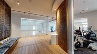 This single-floor fitness facility is thoughtfully designed to accommodate a wide range of athletic and wellness activities. At its core is a full indoor basketball court with soaring ceilings, classic hardwood flooring with regulation court markings, wall-mounted basketball hoops, and protective padding along the walls. High clerestory windows allow natural light to filter into the court space. The court is enclosed by a sleek wire mesh and glass partition system, separating it from the surrounding fitness areas while maintaining visual openness.

Adjacent to the court, the strength and conditioning zone features an extensive array of weight machines, cable systems, rowing equipment, and a heavy punching bag, all set on warm hardwood flooring beneath recessed and track lighting. Full-length mirrors amplify the sense of space. A curved cardio room with large wraparound windows offers outdoor views and houses a row of treadmills, ellipticals, and other aerobic equipment on textured carpet.

A dedicated studio room includes a ceiling-mounted projector, projection screen, spin bikes, stability balls, kettlebells, and floor mats for group or guided workouts. A serene lounge area features low platform seating and a striking decorative laser-cut metal screen wall, providing a space for stretching or recovery. A textured wood accent wall adds warmth near the facility entrance.
