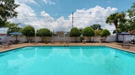 Located in Metairie, Louisiana, this outdoor pool area serves as a central amenity within a two-story residential complex. The centerpiece is a generously sized rectangular swimming pool with clear, vibrant turquoise water, surrounded by a spacious concrete and aggregate deck that provides ample room for lounging and socializing. Multiple seating arrangements are distributed around the pool deck, including wrought iron chairs and tables shaded by blue market umbrellas, offering comfortable spots for relaxation in the Louisiana sun.

The pool area is enclosed by a white privacy fence that defines the perimeter and creates a sense of seclusion, while mature, neatly manicured topiary trees and tropical palms add lush greenery and visual interest throughout the space. Potted plantings punctuate the deck, enhancing the garden-like atmosphere.

Access to the pool area is controlled through ornamental black iron gates with decorative circular detailing, providing both security and an elegant aesthetic. The two-story building facades, clad in light-toned siding and featuring covered balconies, frame the courtyard on multiple sides. Vibrant crape myrtle trees in full pink bloom add seasonal color along the building's entry walkways. A small outbuilding adjacent to the pool area appears to provide additional utility for the space. The overall environment is bright, open, and inviting.