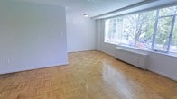 This single-floor space features a clean, minimalist aesthetic defined by its classic parquet hardwood flooring, which adds warmth and character throughout. The flooring is laid in a traditional basket-weave pattern, lending a timeless quality to the interior. The walls are finished in a crisp, neutral white, creating a bright and airy atmosphere that feels open and welcoming.

A large window dominates one wall, fitted with horizontal blinds and framing a view of mature trees outside. This generous window allows abundant natural light to fill the space, enhancing the overall sense of openness. Below the window, a baseboard radiator provides a practical heating solution integrated seamlessly into the room.

The space is open and unobstructed, offering flexibility for a variety of furniture arrangements and uses. A flush ceiling light fixture provides additional illumination, complementing the natural light. The white baseboards throughout neatly frame the parquet floors, adding a polished finishing touch. Overall, the space presents a straightforward, functional layout with enduring architectural details that give it lasting appeal.