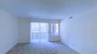 This single-floor space presents a clean, minimalist aesthetic defined by its bright, white walls and light-toned carpeted flooring. The main living area is the focal point of the space, characterized by an abundance of natural light streaming in through a large sliding glass door fitted with vertical blinds, which likely leads to an outdoor area. Adjacent to the sliding door, a secondary window further enhances the room's brightness and ventilation. The white walls and smooth ceiling contribute to an open, airy atmosphere, while the neutral carpet adds warmth underfoot. A wall-mounted HVAC vent is positioned near the ceiling, ensuring climate control throughout. The space's simple, uncluttered design creates a versatile canvas well-suited for a variety of interior arrangements. Overall, the space exudes a fresh, move-in-ready quality, with its light-filled interior and clean finishes offering a welcoming and functional environment.