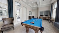This single-floor community lounge space combines leisure and functionality within a refined, coastal-inspired interior. The main area is anchored by a full-size billiards table with a vivid blue felt surface and ornate wooden legs, positioned beneath an elegant coffered ceiling with recessed lighting that adds architectural depth and visual interest overhead.

The space features neutral warm-gray walls complemented by crisp white crown molding and shiplap-style interior doors, creating a polished yet relaxed atmosphere. Plaid-patterned carpeting in soft gray tones runs throughout, tying together the various zones of the room.

Large windows with plantation shutters and floor-to-ceiling navy blue drapes line one wall of the billiards area, inviting generous natural light and offering views of mature outdoor greenery. An adjacent seating nook features a pair of gray leather wingback chairs arranged around a woven accent table, accompanied by a nautical-themed wall painting that reinforces the coastal aesthetic.

A dedicated workstation alcove is built into one wall, housing a wood-toned credenza with computers, a printer, and woven seating—providing a practical workspace within the lounge setting. A wall-mounted television and high-top bistro tables round out the amenities, making this a versatile and well-appointed gathering space.