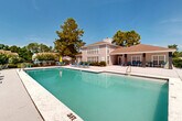 Located at 15235 O'Neal Rd in Gulfport, Mississippi, this outdoor amenity space centers around a generously sized swimming pool with crystal-clear turquoise water, designed to accommodate both leisurely swimming and relaxation. The pool deck is finished in smooth concrete and surrounds the pool on all sides, providing ample space for sunbathing and social gatherings. Numerous lounge chairs and seating arrangements are positioned throughout the deck, offering residents multiple options for outdoor enjoyment.

Adjacent to the pool, a covered patio area extends from the main building, supported by sturdy columns and featuring additional seating beneath its shade. A pergola structure nearby adds an architectural accent to the outdoor space and provides a secondary shaded gathering spot. The main building, finished in neutral gray siding with a pitched shingled roof and large windows, overlooks the pool area and serves as the community's focal point.

The entire pool area is enclosed by a wrought iron fence, ensuring security while maintaining open sightlines to the surrounding landscape. Mature pine and oak trees frame the perimeter, offering natural shade and a lush backdrop. The space benefits from abundant natural sunlight and clear Gulf Coast skies, creating a bright and inviting outdoor environment ideal for year-round recreation.