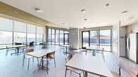 Located at 733 N Drennan St in Houston, TX, this contemporary single-floor space offers a bright and functional layout designed for communal use. The main gathering area is expansive and open, characterized by large floor-to-ceiling windows and glass doors lining multiple walls, flooding the interior with abundant natural light and offering views of the surrounding outdoor landscape. The sleek, large-format gray tile flooring runs throughout, providing a clean and cohesive aesthetic underfoot.

A standout architectural feature is the warm natural wood slat ceiling, which adds texture and visual warmth to the otherwise crisp, modern interior. Recessed lighting complements the natural light, ensuring the space is well-illuminated at all times. The open dining area accommodates multiple table-and-chair groupings, making it well-suited for gathering, dining, or collaborative use.

The kitchen is thoughtfully designed with light wood cabinetry, white countertops, a stainless steel refrigerator, a built-in microwave, and a sink — all finished in a clean, modern style. The kitchen opens seamlessly to the main area, supporting a functional flow throughout the space. Roller shades on the windows offer flexible light and privacy control. Overall, the space balances contemporary design with practical amenities in a welcoming, light-filled environment.
