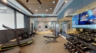 Located at 255 S Mariposa Ave in Los Angeles, this single-floor fitness center offers a well-appointed, contemporary workout environment designed for both functionality and comfort. The space is thoughtfully divided into distinct zones, seamlessly connected through glass partitions that create an open, airy feel while maintaining visual separation between areas.

The main workout area features carpet flooring and is equipped with a comprehensive selection of exercise machines, including IN10CT curved treadmills, air bikes, a dual cable functional trainer, adjustable weight benches, and a full rack of dumbbells. A large wall-mounted mirror runs along one side, enhancing the sense of space and allowing users to monitor their form. A mounted television adds entertainment options during workouts.

Overhead, a distinctive wood-slat ceiling with recessed and track lighting adds warmth and architectural interest, complemented by ceiling fans that aid ventilation. Floor-to-ceiling windows along the perimeter flood the space with natural light and offer views of the surrounding greenery and streetscape.

Adjacent to the gym floor is a bright, welcoming lounge area finished with light hardwood floors, neutral-toned seating, and understated décor, providing a comfortable space to relax before or after a workout. An elevator is also accessible from this lounge area.