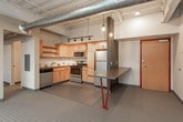 This single-floor loft-style unit showcases a compelling blend of industrial and modern design elements throughout. Exposed concrete ceilings and visible ductwork lend an authentic urban character to the space, while large floor-to-ceiling windows flood the interior with natural light and offer sweeping elevated views of the surrounding landscape and cityscape below.

The kitchen is a functional and well-appointed space, featuring light maple cabinetry with ample upper and lower storage, stainless steel appliances including a refrigerator, range, microwave, and dishwasher, complemented by a stainless steel countertop and a tiled backsplash. A built-in counter with bold red hairpin legs serves as a casual dining or workspace area just adjacent to the kitchen, enhanced by pendant lighting overhead. Track lighting runs along the ceiling, providing flexible illumination throughout.

The bathroom is clean and efficiently designed, with a wood-toned vanity, dark countertop, and well-lit mirror. A stacked washer and dryer unit is conveniently integrated into the bathroom, adding in-unit laundry functionality. Polished concrete floors carry through the space, reinforcing the industrial character while remaining easy to maintain. The overall ambiance is modern, open, and distinctly loft-inspired.