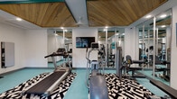 This single-level fitness center is thoughtfully designed to support a comprehensive workout experience. The space features a striking architectural ceiling detail with warm, natural wood panels trimmed in teal, adding a modern and inviting aesthetic to the room. Recessed lighting throughout ensures the space is well-lit, while floor-to-ceiling mirrors along one wall amplify the sense of openness and allow users to monitor their form during exercise.

The fitness center is outfitted with a versatile range of equipment, including a treadmill, elliptical trainer, cable crossover strength machine, adjustable weight bench, and a seated resistance machine. A wall-mounted flat-screen television provides entertainment during workouts. For free weight training, a full rack of dumbbells is available alongside stability balls, catering to a variety of fitness routines.

The flooring consists of a vibrant teal surface accented by bold black-and-white geometric-patterned area rugs, contributing to the space's energetic and contemporary feel. Large windows along one wall frame views of a nearby outdoor pool and mature trees, bringing in natural light and a refreshing outdoor connection. The overall ambiance is clean, modern, and motivating, making it a functional and visually engaging environment for fitness.