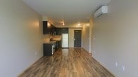 This single-floor unit offers a thoughtfully designed open-concept layout that seamlessly integrates the kitchen and living areas into one cohesive space. The kitchen is outfitted with dark-toned cabinetry that provides ample storage, a stainless steel sink with countertop workspace, a gas range with oven, and a full-size refrigerator. Track lighting above the kitchen adds focused illumination, complementing the flush-mount ceiling fixture that brightens the main living area. Rich, warm-toned hardwood-style flooring extends throughout the entire space, lending a sense of continuity and warmth to the interior. A wall-mounted mini-split air conditioning unit ensures efficient climate control year-round. Neutral wall tones create a clean, versatile backdrop well-suited for a variety of interior styles. A paneled entry door with a classic raised design serves as a welcoming focal point at the far end of the space, with an additional hallway visible beyond, suggesting private rooms or storage tucked away from the main living area. The overall ambiance is modern, functional, and move-in ready.