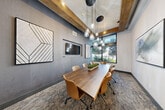 Located at 1050 Blanche St in West Palm Beach, FL, this thoughtfully designed conference room offers a polished and professional environment ideal for meetings and collaborative work. The space is anchored by a substantial walnut wood conference table surrounded by leather upholstered rolling chairs, comfortably seating up to eight. Overhead, a cluster of glass pendant lights with brass accents hangs from a tray ceiling adorned with rich, rustic wood beam detailing, creating a warm contrast against the cool gray-toned walls. A wall-mounted flat-screen television serves as a focal point on one side of the room, flanked by geometric framed art panels that add a contemporary design touch. On the opposite end, a large abstract painting in soft, muted tones adds an artistic element to the bright white wall. Natural light pours in through oversized black-framed windows, offering views of lush tropical greenery that reflect the Florida setting. Patterned carpeting in neutral tones grounds the space and complements the overall aesthetic. The blend of modern finishes and warm natural materials gives this conference room a refined yet inviting ambiance suited for professional gatherings.
