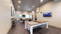 This single-floor community game room is thoughtfully designed to accommodate a wide range of recreational activities in a stylish, welcoming environment. The space is anchored by multiple billiards tables with a clean, modern aesthetic — featuring white and wood-toned finishes — alongside a full-size air hockey table, making it ideal for friendly competition. Overhead, a mix of pendant and chandelier-style light fixtures, including an industrial pulley-style pendant and a ring chandelier with exposed bulbs, add character and warmth to the room.

A dedicated alcove houses a curated arcade corner, complete with classic upright arcade cabinets and an illuminated tabletop gaming console, offering a retro gaming experience within the contemporary setting. Throughout the main area, woven rattan lounge chairs, patterned area rugs, and natural wood accent tables create inviting conversation nooks. A large wall-mounted chalkboard display adds a playful, communal touch.

Wall-mounted flat-screen televisions are positioned throughout for viewing convenience, while bold abstract artwork punctuates the neutral-toned walls. Large windows allow natural light to filter in, and direct exterior access provides a seamless connection to the outdoors. Dark tile flooring ties the entire space together with a polished, cohesive finish.