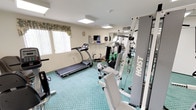 Cypress Park Senior Community is a multi-story residential community distinguished by its classic white exterior, green accent trim, and columned entrance portico. The building's welcoming facade sets the tone for a thoughtfully designed community with a variety of amenities.

The fitness center is a well-appointed space featuring teal patterned carpeting and bright flush-mounted ceiling lights. Residents have access to a diverse selection of equipment, including treadmills, stationary and recumbent bicycles, a Hoist multi-station cable machine, exercise balls, and resistance tools. Full-length mirrors expand the sense of space, and a wall-mounted television provides entertainment during workouts. Natural light filters in through curtained windows, and decorative floral wallpaper adds a warm, inviting touch to one section of the room.

The outdoor areas are equally impressive. A charming courtyard features a decorative pond with natural stonework, lush plantings, and a covered gazebo with a distinctive cupola roof — an ideal setting for relaxation. The expansive community pool area offers a large, curved swimming pool surrounded by a spacious deck furnished with lounge chairs, tables, and umbrellas, all framed by mature trees and the multi-story residential buildings above.