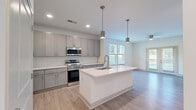 This well-designed one-bedroom, one-bathroom unit in Carmel, Indiana offers a thoughtfully arranged floor plan that maximizes its efficient footprint. The open-concept kitchen and living area serves as the heart of the space, where light wood-tone flooring flows seamlessly throughout. The kitchen is appointed with grey shaker-style cabinetry, a white quartz countertop island with an undermount sink, stainless steel appliances including a refrigerator, range, and over-the-range microwave, and a classic white subway tile backsplash. Pendant lighting above the island adds a modern touch, while recessed lighting ensures the space remains bright. The living room features a coffered accent wall with decorative paneling and access to an outdoor area via a glass door, filling the room with natural light. The bedroom is carpeted, offers a ceiling fan, and benefits from windows overlooking outdoor greenery. The bathroom is finished with grey cabinetry, a quartz vanity, pendant lighting, a large mirror, and light wood-tone flooring. A walk-in closet equipped with wire shelving and carpeted flooring provides practical storage.