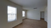 This single-floor space presents a clean, straightforward layout centered around a spacious main room finished in a neutral palette. The walls are painted a soft white, contributing to a bright and airy atmosphere that is further enhanced by two windows fitted with white horizontal blinds. These windows allow natural light to filter in and offer views of the surrounding neighborhood exterior. The room features wall-to-wall beige carpeting, providing a warm and comfortable underfoot feel throughout the space. The ceiling is smooth and white, housing a centrally positioned light fixture and a ceiling-mounted HVAC vent, indicating a forced-air climate control system. Baseboard trim runs along the perimeter of the room, adding a clean, finished look to the space. An additional wall-mounted vent is visible near the upper corner of the room, suggesting efficient air circulation. Electrical outlets are positioned at standard intervals along the walls, and a light switch or outlet panel is visible on one wall. The overall ambiance is minimal and neutral, making it a versatile blank canvas well-suited for a variety of uses and personal customization.