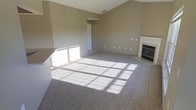 This single-floor space presents an open and airy living area characterized by vaulted ceilings that enhance the sense of volume and spaciousness. The room is finished with neutral, warm-toned walls that complement the plush, light-colored carpeting throughout. A white-mantled fireplace anchors one wall, serving as a focal point and adding a classic architectural detail to the space. Large windows along one wall allow generous amounts of natural light to flood the interior, casting bright, grid-patterned shadows across the floor and contributing to an inviting atmosphere. An adjacent doorway leads to additional areas of the home, suggesting a functional layout with clear transitions between spaces. The open floor plan offers flexibility for various furniture arrangements, while the clean, move-in-ready condition of the space highlights its well-maintained finishes. The combination of vaulted ceilings, natural light, soft carpeting, and the fireplace creates a comfortable and versatile living environment suitable for everyday use.