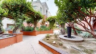 Located at 102 N Lemon Ave in Ontario, California, this outdoor courtyard space showcases a thoughtfully designed Mediterranean-inspired environment that blends architecture and landscaping into a cohesive, inviting retreat. The building exterior features a warm terracotta and stucco façade accented by arched doorways, balconies with iron railings, and a pergola draped in climbing vines, all contributing to a classic, refined aesthetic.

The courtyard is defined by raised planters with brick-capped terracotta retaining walls that organize a rich variety of plantings, including ornamental grasses, agave, cacti, flowering shrubs, and mature shade trees. A central lawn area provides open green space, while paved concrete pathways guide visitors through the landscape. A decorative water fountain serves as a focal point, adding a tranquil, resort-like atmosphere to the setting.

Palm trees punctuate the perimeter, reinforcing the Southern California character of the space. Bollard lighting and classic lamp posts are positioned throughout to ensure the courtyard remains functional and attractive after dark. The overall ambiance is serene and well-maintained, offering residents and visitors a picturesque outdoor environment that balances natural elements with architectural detail.
