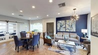 This single-story space showcases an open-concept layout that seamlessly connects the living and dining areas, creating a cohesive and functional environment. The living area is anchored by a bold navy accent wall adorned with a large-scale abstract triptych in swirling blues, greens, and yellows, serving as a dramatic focal point. A grey sofa with striped accent pillows and a marble-top coffee table furnish the space, complemented by an elegant globe chandelier overhead. Recessed lighting throughout ensures consistent, well-distributed illumination across the entire area. Adjacent to the living space, the dining area features a round dark-wood table surrounded by upholstered blue chairs, positioned near an arched window that allows natural light to filter in and offers views of the surrounding greenery. Wood-look tile flooring and a patterned area rug add warmth and texture underfoot. A louvered interior door and neutral grey walls balance the bolder design elements, contributing to a polished yet welcoming ambiance throughout the space.