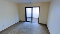 This single-floor space features a generously sized room finished with wall-to-wall carpet in a neutral tone, providing a comfortable and practical foundation underfoot. The walls are painted in a soft, warm off-white, contributing to a bright and clean aesthetic throughout. A standout feature of the room is the sliding glass door framed in dark wood trim, which serves as both a functional entry point to an adjoining outdoor balcony or patio and a significant source of natural light. The exterior space visible through the glass appears to be an enclosed or semi-enclosed balcony, adding an additional layer of usable square footage and offering outdoor views. The room's layout is open and unobstructed, making it highly versatile for various uses. Simple white baseboards run along the perimeter of the room, complementing the understated color palette. The overall ambiance is clean, neutral, and move-in ready, with an emphasis on practicality and natural illumination. The combination of soft carpeting, light-toned walls, and the glass door access to the exterior creates a functional and welcoming environment.
