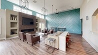 Located at 427 Betty Ln in Ewing, NJ, this single-floor community amenity facility offers a thoughtfully designed layout across a generous footprint. The centerpiece is a dramatic living room featuring soaring double-height ceilings adorned with bold teal herringbone-patterned accent walls and a row of clerestory windows that flood the space with natural light. A built-in fireplace with flanking shelving units anchors the room, complemented by stylish lantern-style pendant lighting and durable wood-look flooring throughout. The adjacent combined dining and living area transitions seamlessly from the main lounge, offering a tall communal dining table with bar-height seating, pendant lighting, and large windows overlooking lush outdoor greenery. A cozy game nook features a round poker-style table surrounded by upholstered chairs beneath an elegant chandelier, bathed in natural light from wraparound windows. The exercise room is expansive and well-appointed, housing a full array of cardio machines including treadmills, ellipticals, and stationary bikes, alongside strength equipment. Full-length mirrored walls enhance the sense of space, while a dedicated yoga and stretching studio features motivational wall murals, light hardwood floors, built-in shelving, and serene garden views.