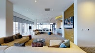 This single-floor space presents a thoughtfully designed open-plan lounge area that combines modern aesthetics with functional comfort. The expansive layout is anchored by large floor-to-ceiling windows along the perimeter, flooding the interior with abundant natural light while offering expansive urban views of the surrounding cityscape.

The main lounge area features multiple seating arrangements, including a generous sectional sofa paired with accent chairs in warm yellow tones and complementary cushions in teal and neutral shades. A sleek, dark coffee table serves as a central focal point, while a textured area rug defines the primary seating zone. Additional seating clusters with modular sofas and a teal ottoman are positioned toward the window-lined walls, creating distinct conversation areas within the open space.

A wood-paneled accent wall houses a large flat-screen television, adding warmth and visual contrast against the white ceilings and walls. Recessed lighting throughout the ceiling provides consistent, even illumination, supplemented by a tall floor lamp. The smooth transition of hardwood flooring and carpet adds textural variety. The overall ambiance is contemporary, bright, and welcoming.