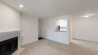 This single-floor space offers a clean, modern aesthetic defined by crisp white walls, smooth ceilings, and neutral wall-to-wall carpeting that creates a warm and cohesive feel. The open-concept living area is airy and well-lit, providing a versatile canvas for a variety of furniture arrangements. A defining architectural feature is the pass-through kitchen window, which connects the kitchen to the living area, enhancing the sense of openness while maintaining a defined cooking space. The kitchen is outfitted with white cabinetry, a stainless steel refrigerator, a sink, and dark countertops — offering a functional and stylish workspace. A doorway visible from the main living area leads to additional spaces, suggesting a practical and efficient floor plan. The unit's interior palette of whites and soft neutrals maximizes the perception of light and space throughout. Recessed lighting in the kitchen and flush ceiling fixtures contribute to a consistently bright atmosphere. The thoughtful layout and minimalist design make this a highly functional space with a tidy, contemporary character suited for comfortable everyday living.