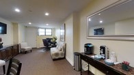 This thoughtfully designed single-floor space offers a welcoming and functional environment suited for both work and leisure. The interior is finished with warm, neutral tones — soft cream walls and plush carpeted flooring — creating a calm and professional ambiance throughout.

The main area is divided into distinct functional zones. A lounge area features a stylishly patterned accent chair, providing a comfortable spot for relaxation or informal meetings. Adjacent to this, a dedicated workspace is furnished with a desk and ergonomic office chair, positioned near a window that allows natural light to filter in, enhancing the productivity of the area.

A console table along one wall serves as a refreshment station, equipped with a single-serve coffee maker and additional beverage accessories, all set atop a rich, dark granite surface. A large framed mirror mounted above the console adds depth and brightness to the space. Recessed ceiling lighting is distributed evenly throughout, ensuring a well-lit interior regardless of natural light availability.

Decorative touches, including fresh floral arrangements and framed wall art, add character and warmth to the space. A sleek stainless steel waste bin completes the tidy, organized aesthetic. Overall, this space balances practicality with comfort in a clean, polished setting.