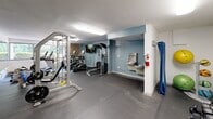 This fitness center at 600 Martin Luther King Jr Blvd in Chapel Hill, NC offers a well-equipped exercise space designed to support a full range of workout routines. The room features durable rubber flooring throughout, providing a safe and practical surface for both cardio and strength training activities. Natural light filters in through a row of windows along one wall, offering views of the surrounding greenery and keeping the space bright and inviting. Exposed brick accents add a touch of warmth to the otherwise clean, modern interior with white and soft blue walls. The cardio section is equipped with multiple treadmills and elliptical machines, while a multi-station cable machine and a barbell rack with weight plates cater to strength training needs. A dumbbell rack stocked with a variety of free weights, along with exercise balls, provides additional versatility. A large wall mirror enhances the sense of space and allows users to monitor their form. A wall-mounted flat-screen TV adds entertainment options during workouts. The room also includes a dual water fountain station for convenient hydration and a fire extinguisher for safety. Overhead fluorescent lighting ensures the space remains well-lit at all times, complementing the natural light from the windows.