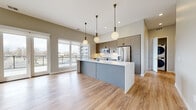 This single-floor residence showcases a thoughtfully designed open-concept layout with a contemporary aesthetic. Light wood-tone flooring flows continuously through the main living areas, enhancing the sense of openness and cohesion throughout the space.

The kitchen serves as a functional centerpiece, featuring a large island with a crisp white countertop and integrated sink, complemented by two-toned cabinetry in taupe and slate gray with matte black hardware. A white subway tile backsplash, stainless steel appliances—including a gas range, built-in microwave, and French door refrigerator—and globe pendant lights with brass accents complete the well-appointed kitchen. Large windows flood the area with natural light and offer views of the surrounding urban landscape.

The open living area extends from the kitchen, featuring French doors and expansive windows that lead to an outdoor balcony, filling the space with additional natural light. A secondary bedroom provides hardwood flooring and a generous window with exterior views.

The bathroom is finished with white stacked tile surrounding a soaking tub and shower combination, chrome fixtures, stone-look tile flooring, and ample built-in cabinetry with black hardware. A dedicated walk-in closet offers customizable shelving, hanging rails, and pull-out wire baskets, providing generous storage. The unit is accessed through a solid wood entry door with a keypad lock.