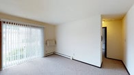 This single-floor residential space offers a clean, functional layout designed for comfortable everyday living. The main living area is finished with neutral-toned carpet and features crisp white walls that create a bright, open atmosphere. Large sliding glass doors dressed with vertical blinds allow an abundance of natural light to fill the room while providing access to the outdoors and views of surrounding greenery. A baseboard heating system runs along the perimeter of the living area, ensuring efficient climate control throughout the space. The room's simple, understated design provides a versatile canvas that adapts easily to a variety of furnishing styles. A partial wall divides the living area from the adjoining spaces, contributing to a sense of structure without compromising the open feel of the floor plan. A glimpse into the adjacent area reveals a tiled or hard-surface floor and what appears to be a kitchen or utility space, suggesting a practical and well-organized layout. The overall ambiance is light, neutral, and move-in ready, making this a straightforward and welcoming residential space suited for a range of occupants.