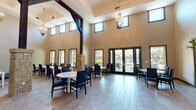 This well-appointed community space spans a generous single floor, thoughtfully designed to serve as a social and gathering hub. The main hall is defined by dramatically soaring ceilings that create an open, airy atmosphere, accentuated by a row of clerestory windows along the upper walls that flood the interior with natural light throughout the day. Dark wood exposed beams and a stone column accent add warmth and architectural character to the neutral, beige-toned walls.

The primary gathering area features light-toned tile flooring and is furnished with multiple round and rectangular white tables paired with navy blue upholstered chairs, providing flexible seating arrangements. Elegant pendant chandeliers hang overhead, enhancing the refined ambiance. Large glass French doors open directly to an outdoor patio, offering views of greenery beyond.

A secondary dining or lounge area provides additional seating along windowed walls, while a separate room features a wall-mounted flat-screen television for media viewing. The kitchenette is a standout feature, showcasing a bold black-and-white patterned mosaic tile backsplash, marble-style countertops, light cabinetry, and pendant lighting — ideal for food preparation and service. Framed artwork accents the transitional spaces, completing the polished, community-focused design.