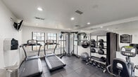 Located at 1810 W Apollo Rd in Garland, TX, this single-story fitness center offers a well-equipped workout facility designed to support a range of exercise routines. The space is organized into distinct functional zones, making efficient use of its layout.

The main workout area features multiple treadmills, including SportsArt models, positioned along windows that provide natural light and views of an outdoor pool area. A Vision dual cable machine serves as a centerpiece for strength training, complemented by a large wall mirror that visually expands the space. A dedicated free weights zone includes a multi-tiered rack stocked with medicine balls and stability balls, along with a set of kettlebells for functional training.

A secondary area houses additional cardio equipment, including a rowing machine and a stationary bike, alongside a wall-mounted flat-screen television for entertainment during workouts. A wall-mounted water fountain and waste receptacle contribute to the facility's practical amenities. Posted fitness center rules provide guidance for users.

Throughout the space, dark rubber tile flooring offers durability and traction, while recessed lighting and white walls create a clean, bright environment. Multiple windows ensure consistent natural light, contributing to an open and energizing atmosphere.