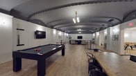 This single-floor space is defined by its most prominent architectural feature: a dramatic barrel-vaulted ceiling finished in deep gray, adorned with detailed ornate crown molding that runs along the curved perimeter. The contrast between the dark ceiling and crisp white walls creates a bold, sophisticated atmosphere throughout. Light wood flooring extends across the entire space, lending warmth and cohesion to the various activity zones within.

The lounge area is furnished with a generous arrangement of low-profile dark sofas and accent chairs, centered around a wall-mounted flat-screen television. Warm sconce lighting punctuates the white walls, complemented by a modern pendant chandelier overhead. A sideboard and decorative accessories add refined finishing touches to this relaxed seating zone.

Adjacent to the lounge, an open recreation area houses a full-size pool table and high-top tables with bar stools, creating an inviting social atmosphere. Framed artwork is displayed throughout, adding a curated aesthetic to the space.

A dedicated game room, clearly signed, features a foosball table, ping-pong table, and additional seating. This area benefits from clean, bright flush-mount lighting and a more casual, energetic feel. Together, the zones form a well-rounded, multi-use venue suited for socializing and recreation.