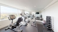 This fitness center offers a comprehensive workout environment designed to support a wide range of exercise routines. The space is finished with durable speckled rubber flooring throughout, providing both safety and functionality underfoot. Crisp white walls and recessed lighting keep the interior feeling bright and open, while a large wall-mounted mirror along one side enhances the sense of depth and allows users to monitor their form during workouts.

Natural light floods the space through multiple large windows that frame sweeping elevated views of the surrounding treetop landscape, creating an energizing atmosphere. The cardio area is well-stocked with a lineup of elliptical trainers, upright and recumbent stationary bikes, and additional machines suited for low-impact exercise. A cable functional trainer and a weight bench with a dumbbell rack provide options for strength and resistance training, complemented by stability balls and suspension training straps mounted to the wall.

A wall-mounted flat-screen television is positioned for easy viewing during workouts, and a white storage cabinet with louvered paneling provides a tidy station for equipment and supplies. Instructional workout posters are displayed on the walls for reference. A glass-paneled door leads to an adjacent area, adding connectivity to the overall facility. The space is clean, organized, and thoughtfully arranged to accommodate multiple users simultaneously.