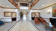 This distinguished commercial space makes a strong first impression through its grand entrance lobby, which showcases soaring double-height ceilings crowned by a sweeping curved mezzanine balcony with rich wood railings and decorative metal balusters. The flooring throughout the main corridor is finished in polished marble-style tile laid in a classic geometric pattern with contrasting dark diamond inlays, lending an air of sophistication and formality to the space.

The reception area is positioned to the right of the main corridor and features warm wood-trimmed furnishings, a traditional desk setup, and comfortable upholstered seating in deep red tones — ideal for welcoming guests and visitors. Crown molding runs along the upper walls, reinforcing the refined, traditional aesthetic carried throughout the space.

The long central hallway extends toward a set of glass entry doors at the far end, allowing natural light to filter deep into the interior. Recessed lighting and classic ceiling-mounted fixtures provide consistent, warm illumination throughout. Additional office or service areas are accessible from the main corridor, visible through wood-framed doorways on both sides. Overall, the space conveys a professional, formal ambiance suited to an institutional, medical, or corporate setting.