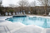 This outdoor pool area offers a well-designed recreational retreat set against a backdrop of mature trees and open skies. The centerpiece is a generously sized freeform swimming pool with clear, blue-tinted water and a smooth concrete surround that provides ample space for lounging and movement. Depth markers are clearly indicated, with shallow entry points and a gradual transition to deeper sections, making the pool accessible for a range of swimmers.

The expansive pool deck is finished in a light-toned concrete surface that reflects natural light and remains comfortable underfoot. A collection of sling-style lounge chairs lines the perimeter, complemented by shaded umbrella stations that offer relief from direct sun. The layout encourages both active use and relaxed sunbathing.

A gated entry with clearly posted pool rules provides a secure and organized point of access. The surrounding wrought iron fence encloses the entire area, ensuring safety while maintaining open sightlines to the natural landscape beyond. Adjacent to the pool, a classic clubhouse structure with a covered porch and French doors serves as a functional complement to the outdoor space. Pergola accents and manicured pathways further enhance the overall aesthetic, creating a cohesive and inviting community amenity space.