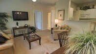 This single-floor residence centers around a living room finished in warm, neutral tones that create a calm and inviting atmosphere. The space is carpeted throughout, adding a soft, comfortable feel underfoot. The living area is furnished with a plush sofa accented by decorative throw pillows, a classic wooden coffee table with a glass top, and a matching accent chair, all arranged to encourage conversation and relaxation. A wall-mounted flat-screen television is positioned opposite the seating area, complemented by a console table below displaying decorative accents, including a traditional mantel clock. A framed art piece adorns one wall, while a table lamp provides warm ambient lighting. The open layout allows clear sightlines to interior doorways leading to additional rooms, suggesting a practical and connected floor plan. A pass-through or shelf detail near one wall hints at a nearby kitchen or dining area. The overall design aesthetic leans traditional, with dark wood furniture contrasting against the light-colored walls and carpet, creating a cohesive and well-appointed interior that balances functionality with classic style.