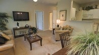 This single-floor residence centers around a living room finished in warm, neutral tones that create a calm and inviting atmosphere. The space is carpeted throughout, adding a soft, comfortable feel underfoot. The living area is furnished with a plush sofa accented by decorative throw pillows, a classic wooden coffee table with a glass top, and a matching accent chair, all arranged to encourage conversation and relaxation. A wall-mounted flat-screen television is positioned opposite the seating area, complemented by a console table below displaying decorative accents, including a traditional mantel clock. A framed art piece adorns one wall, while a table lamp provides warm ambient lighting. The open layout allows clear sightlines to interior doorways leading to additional rooms, suggesting a practical and connected floor plan. A pass-through or shelf detail near one wall hints at a nearby kitchen or dining area. The overall design aesthetic leans traditional, with dark wood furniture contrasting against the light-colored walls and carpet, creating a cohesive and well-appointed interior that balances functionality with classic style.