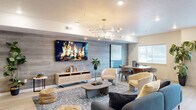 This single-floor community space showcases a thoughtfully designed open-concept layout that seamlessly connects a fully equipped kitchen, a lounge area, and a dining zone into one cohesive, welcoming environment. The kitchen is appointed with white shaker-style cabinetry, open display shelving with subway tile backsplash, stainless steel appliances including a full-size refrigerator and microwave, and a spacious island with a quartz countertop and bar seating. Light wood-look flooring flows throughout the entire space, complementing the warm, contemporary aesthetic. A striking accent wall clad in gray wood-look paneling anchors the lounge area and serves as the backdrop for a large wall-mounted television, with a rattan media console below. The lounge features multiple seating arrangements, including plush sofas, curved accent chairs, and a modern sculptural coffee table, all centered on a patterned area rug. A bold sputnik-style chandelier adds a sophisticated focal point overhead. A secondary seating area is painted in a calming muted teal, adorned with large-scale artwork, a decorative cabinet with an arched mirror, and abundant natural light from nearby windows. A round dining table with upholstered chairs provides an additional gathering spot, rounding out this versatile and elegantly appointed shared space.