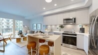 This thoughtfully designed 2-bedroom, 2-bathroom residence on a single floor offers a seamless open-concept layout that maximizes every inch of its compact footprint. The modern kitchen is a standout feature, outfitted with sleek flat-front cabinetry in soft grey tones, a white quartz countertop, subway tile backsplash, and a full suite of stainless steel appliances including a range, microwave, dishwasher, and refrigerator. A light wood island with bar seating provides both additional prep space and a casual dining option. The kitchen flows naturally into the living room, which is bright and airy thanks to large floor-to-ceiling doors that open to a private outdoor area, inviting natural light throughout. Light hardwood flooring runs continuously across the main living areas, enhancing the sense of warmth and cohesion. The primary bedroom is a comfortable retreat featuring a large window with urban views and a sliding closet. The second bedroom is similarly well-proportioned and bright. Both bathrooms are finished in clean whites with modern chrome fixtures — one featuring a combined shower and soaking tub, the other a streamlined vanity with ample counter space. A hallway and closet round out the efficient and functional floor plan.