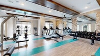 This fitness center combines modern design with a comprehensive selection of exercise equipment, creating a functional and visually appealing workout environment. The space is anchored by a row of treadmills and cardio machines, including stationary bikes and ellipticals, all positioned to take advantage of the natural light streaming in through large windows and sliding glass doors. A full dumbbell rack lines one wall, catering to free-weight training, while a multi-functional cable machine and weight bench occupy a dedicated strength training zone. The flooring features a light wood-look tile accented by distinctive teal-colored runner strips, adding a contemporary aesthetic throughout. Overhead, rich dark wood beams span the white ceiling, lending warmth and architectural character to the space. Ceiling fans are mounted throughout to ensure comfort during workouts. Floor-to-ceiling mirrors extend one wall, enhancing the sense of openness and allowing users to monitor their form. A textured herringbone-patterned accent wall adds a refined design touch. Glass-paneled doors provide connectivity to adjacent areas of the facility, contributing to an open, airy atmosphere. The overall design strikes a balance between upscale aesthetics and practical fitness functionality.