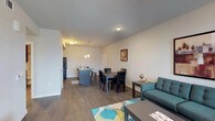 This single-floor unit presents a thoughtfully designed open-concept layout that seamlessly connects the living, dining, and kitchen areas. The living space features warm hardwood-style flooring, neutral walls, and a teal sofa, creating an inviting atmosphere. A dining area accommodates a full-sized table with seating for four, positioned beneath pendant lighting that adds a stylish focal point.

The kitchen is well-equipped with rich dark wood cabinetry, granite countertops, and black appliances, including a range and microwave. A kitchen island with bar seating provides additional prep and dining space, illuminated by pendant lights overhead. The open sightlines from the kitchen into the living area enhance the sense of spaciousness throughout.

The bedroom is carpeted and generously sized, offering multiple closet spaces with double doors and a ceiling fan for added comfort. The bathroom features a granite-topped vanity with dark wood cabinetry, an undermount sink, and a well-lit mirror with a row of modern sconce lighting above.

A separate den or flex space benefits from abundant natural light through large windows fitted with horizontal blinds, with a glass-paneled entry door that further brightens the room, making it well-suited as a home office or reading nook.