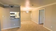 This single-floor residence offers a clean, open-concept design that promotes a sense of spaciousness and easy flow between living areas. The living space is finished with wall-to-wall carpeting in a warm neutral tone, complemented by light beige walls and bright white crown molding and baseboards that add a polished, classic touch throughout.

A decorative pendant light hangs at the center of the dining area, providing focused illumination and serving as a subtle focal point within the open layout. The space benefits from a partially raised kitchen counter that creates a natural visual division between the kitchen and living areas while maintaining an open feel.

The kitchen features white cabinetry, a dark countertop, and includes standard appliances such as a range and refrigerator. A decorative column with crown molding detailing anchors the kitchen's entry point, adding architectural character to the transition between spaces.

A white paneled door leads to an adjoining room, contributing to the unit's functional layout. Ceiling vents are integrated throughout, ensuring consistent climate control. The overall ambiance is bright, neutral, and move-in ready, offering a versatile canvas well-suited for a variety of interior styles.