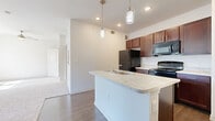This single-floor unit offers a thoughtfully designed layout that maximizes its compact footprint. The open-concept kitchen serves as the heart of the space, showcasing rich dark wood cabinetry, light stone-style countertops, and a full suite of black appliances including a refrigerator, gas range, and built-in microwave. A central kitchen island with a built-in sink and matching countertop provides added workspace and functions as a natural divider between the kitchen and the living area. Elegant pendant lighting hangs above the island, complementing the recessed ceiling lights throughout. Hardwood-style flooring runs through the kitchen and main areas, lending warmth and continuity. The living area transitions to soft, plush carpeting and features large windows with blinds alongside a sliding glass door that invites natural light and outdoor views. Ceiling fans are present to enhance airflow and comfort. The bathroom is finished with tile-surround tub and shower combination, hardwood-style flooring, and a dark wood vanity with a light countertop. A dedicated closet provides practical storage. The overall palette of neutral whites and warm wood tones creates a clean, inviting atmosphere throughout.