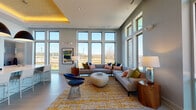 Located at 4011 Tryon Park Rd in Charlotte, NC, this single-floor community clubhouse delivers a sophisticated and welcoming atmosphere through a seamless blend of contemporary design and thoughtful functionality. The space is anchored by a generous open-concept lounge area with dramatically high ceilings and expansive floor-to-ceiling windows on multiple walls, flooding the interior with abundant natural light and offering sweeping views of the surrounding landscape and community grounds. Light-toned large-format tile flooring runs throughout, contributing to the bright, airy character of the space. The lounge is centered around a curved sectional sofa accented with richly colored throw pillows, a sculptural oval ottoman, and a vibrant gold-toned area rug, all set beneath a distinctive tray ceiling detail with warm ambient uplighting. Transitioning into the entertaining zone, a long white quartz island with bar-height seating serves as the focal point, complemented by bold oversized pendant lighting and a wall-mounted television. Built-in display niches with decorative lighting add architectural interest, while a fully equipped wet bar with sink and appliances ensures the space is ready for hosting. A ceiling-mounted projector further enhances the entertainment capabilities of this well-appointed clubhouse.