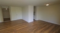This single-floor unit offers a clean, functional layout with a focus on open living. The main living area is spacious and unobstructed, finished with warm-toned hardwood-style flooring that runs throughout the space, lending a cohesive and inviting feel underfoot. The walls are painted in a neutral off-white tone, providing a bright, versatile backdrop suited to a variety of décor styles. A flush-mount ceiling light provides overhead illumination in the living area.

A partial dividing wall separates the living space from the kitchen, creating a degree of definition between the two areas without fully enclosing them. The kitchen features white upper cabinetry and white appliances, including what appears to be a stove, contributing to a clean and utilitarian aesthetic. The entry door, finished in white, is positioned along the main wall of the living area and includes standard hardware and a security panel nearby.

Overall, the space presents a straightforward, move-in-ready layout with consistent flooring, neutral finishes, and a practical kitchen configuration—making it a functional and adaptable living environment.