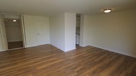 This single-floor unit offers a clean, functional layout with a focus on open living. The main living area is spacious and unobstructed, finished with warm-toned hardwood-style flooring that runs throughout the space, lending a cohesive and inviting feel underfoot. The walls are painted in a neutral off-white tone, providing a bright, versatile backdrop suited to a variety of décor styles. A flush-mount ceiling light provides overhead illumination in the living area.

A partial dividing wall separates the living space from the kitchen, creating a degree of definition between the two areas without fully enclosing them. The kitchen features white upper cabinetry and white appliances, including what appears to be a stove, contributing to a clean and utilitarian aesthetic. The entry door, finished in white, is positioned along the main wall of the living area and includes standard hardware and a security panel nearby.

Overall, the space presents a straightforward, move-in-ready layout with consistent flooring, neutral finishes, and a practical kitchen configuration—making it a functional and adaptable living environment.