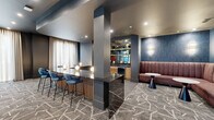 Located at 7800 E Peakview Ave in Centennial, Colorado, this thoughtfully designed lounge space offers a sophisticated, contemporary atmosphere across a single open floor. The layout is anchored by a striking curved bar counter finished in dark marble-veined stone, wrapped in warm wood slatted paneling and lined with vibrant blue velvet barstools — a clear centerpiece for social gatherings. Globe accent lighting along the bar adds an intimate, modern glow.

Adjacent to the bar, a richly upholstered mauve banquette seating area is set against a textured blue accent wall with elegant wall sconces, creating a cozy yet refined nook ideal for conversation. Open shelving and a wet bar station in the background provide functional amenities within easy reach.

The open lounge area features plush dark armchairs grouped around low round tables, offering flexible seating arrangements. Large windows along one wall allow natural light to filter in through sheer drapery, lending an airy quality to the space. Decorative elements, including a sculptural red botanical arrangement and organic wall art, add artistic character throughout.

Underfoot, a bold geometric-patterned carpet in grey and white ties the entire space together, reinforcing the modern design language. Recessed lighting throughout ensures consistent, well-distributed illumination. The overall ambiance is polished, welcoming, and well-suited for both social and professional gatherings.