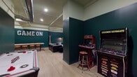 Located at 57 Cannon St in Poughkeepsie, NY, this single-floor entertainment space is thoughtfully designed to inspire fun and social connection. The room is immediately defined by its bold two-tone color scheme — deep forest green on the lower walls transitioning to a soft sage on the upper walls and ceiling — creating an energetic yet cohesive atmosphere throughout.

A bold "GAME ON" marquee sign anchors the main wall, setting the tone for the space. Classic arcade cabinets, including NBA Jam and a Legends multi-game unit, are positioned in one corner alongside a bar-height stool, offering a nostalgic gaming experience. An air hockey table occupies a central area, providing an interactive focal point for groups.

The lounge area features a large sectional sofa, a blue velvet armchair, and a colorful area rug, all arranged to encourage relaxed conversation. A small bistro-style chess table with paired chairs offers a quieter, more intimate game option. A generously sized communal dining table with industrial-style legs and leather chairs provides ample seating for group gatherings or tabletop games, complemented by a well-stocked shelf of board games nearby.

Exposed ductwork and recessed lighting add an industrial character to the ceiling, while light wood flooring ties the entire space together. Freestanding coat racks and thoughtful furniture placement ensure the layout remains open, functional, and welcoming.