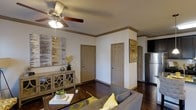 This single-story space offers a well-appointed interior characterized by rich, dark hardwood flooring that extends throughout the main living area. The living room serves as the central hub of the space, featuring a ceiling fan with integrated lighting that provides both functionality and a casual aesthetic. Crown molding with a warm taupe finish adds a refined architectural detail along the upper walls, framing the room with a polished touch.

The walls are painted in a crisp, neutral white, creating a clean backdrop that complements the earthy tones found in the trim, doors, and décor. Two interior doors with raised panel designs and taupe finishes open from the living area, likely leading to private rooms. Abstract wall art in muted gold and grey tones enhances the contemporary feel of the space.

A sideboard-style console table provides storage and display space along one wall, accompanied by decorative accessories. The living area accommodates a grey sofa accented with yellow throw pillows, adding a pop of color. Adjacent to the living room, a partial view of the kitchen reveals a stainless steel refrigerator, suggesting a functional and connected layout throughout.