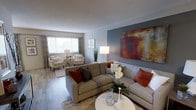 Located at 414 N Forest Park Blvd in Knoxville, Tennessee, this single-floor residence offers a well-designed living space with a contemporary aesthetic and inviting atmosphere. The living area serves as the heart of the home, showcasing a combination of neutral tones and bold accent elements. A striking feature wall in a cool slate-blue hue provides an eye-catching backdrop for a large, richly-colored abstract artwork, adding warmth and visual depth to the space. The room is anchored by a generously sized sectional sofa in a warm taupe tone, accented with rust and charcoal throw pillows that complement the overall color palette. A dedicated sitting nook near the window offers a cozy retreat, furnished with matching accent chairs and a bistro-style side table, bathed in natural light filtering through large windows dressed with patterned drapery and horizontal blinds. The flooring throughout is a durable wood-look material in a medium tone, grounding the space with texture and continuity. Soft area rugs define the seating zones, adding comfort underfoot. The thoughtful layering of lighting, including a tall floor lamp and natural light, ensures the space remains bright and welcoming throughout the day. Overall, the home presents a polished, move-in-ready environment ideal for comfortable everyday living.