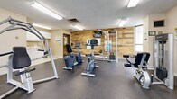 Located at 4 Silver Maple Crt in Brampton, Ontario, this fitness facility offers a well-organized and functional space designed to support a wide range of workout routines. The room features durable rubber flooring throughout the main exercise area, providing a safe and practical surface for equipment use, transitioning to light ceramic tile near the kitchenette zone.

The cardio section is well-appointed with multiple treadmills and a rowing machine, complemented by natural light streaming in through large windows dressed with vertical blinds. A full-length wall mirror enhances the sense of openness and allows users to monitor their form during exercise. The strength training area includes stationary bikes, a cable resistance machine, a dumbbell rack with a flat bench, and additional multi-use equipment, all anchored by a warm wood-paneled accent wall that adds a distinctive design touch.

Overhead fluorescent lighting ensures the space is consistently well-lit throughout. A dedicated kitchenette area along one wall features white cabinetry, a sink, a water cooler, and a coffee maker, offering a convenient station for hydration and refreshment. Safety equipment, including a fire extinguisher, is also present. The overall ambiance is clean, practical, and welcoming, making this fitness room a comprehensive amenity for residents.